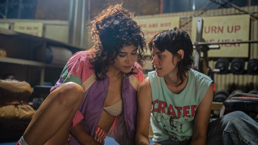 A film still of Katy O'Brian and Kristen Stewart sitting close together, in 80s-style outfits, on a gym floor.