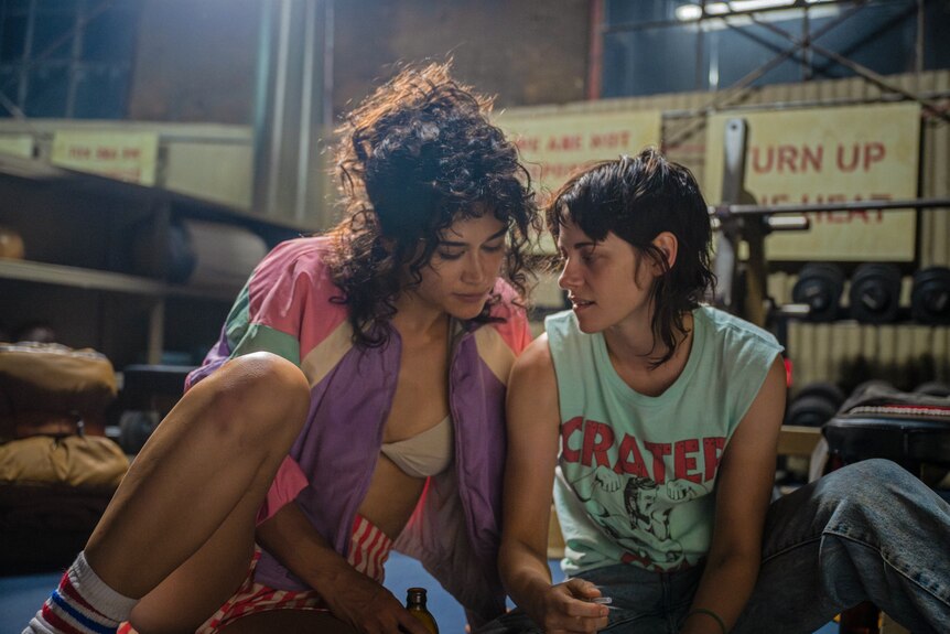 A film still of Katy O'Brian and Kristen Stewart sitting close together, in 80s-style outfits, on a gym floor.
