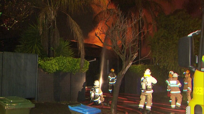 Firefighters standing next to a house fire with one directing a hose pointing water towards it