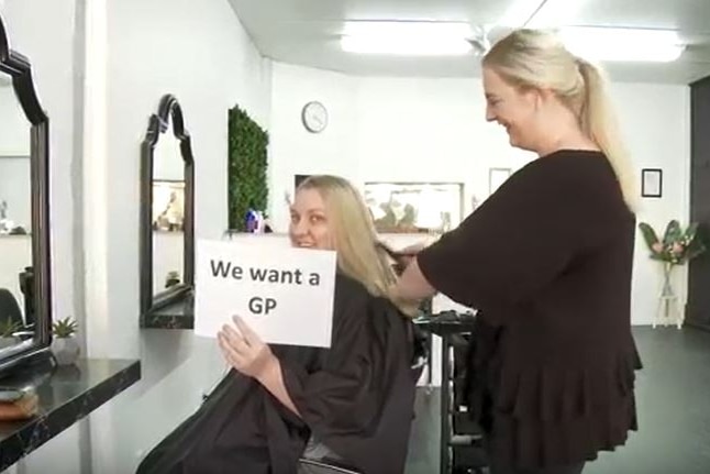 A woman cuts the hair of another woman, who holds a sign saying 'We want a GP'.