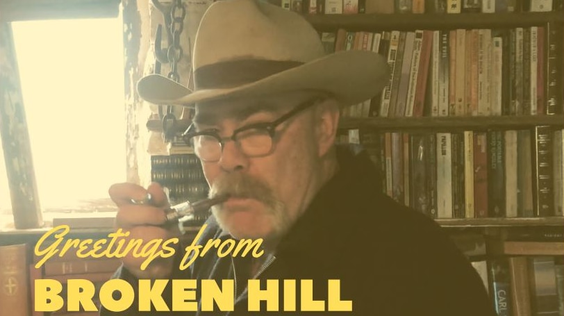 A head shot of a man in a large cowboy hat smoking a pipe standing against a bookcase.  Welcome to Broken Hill is below