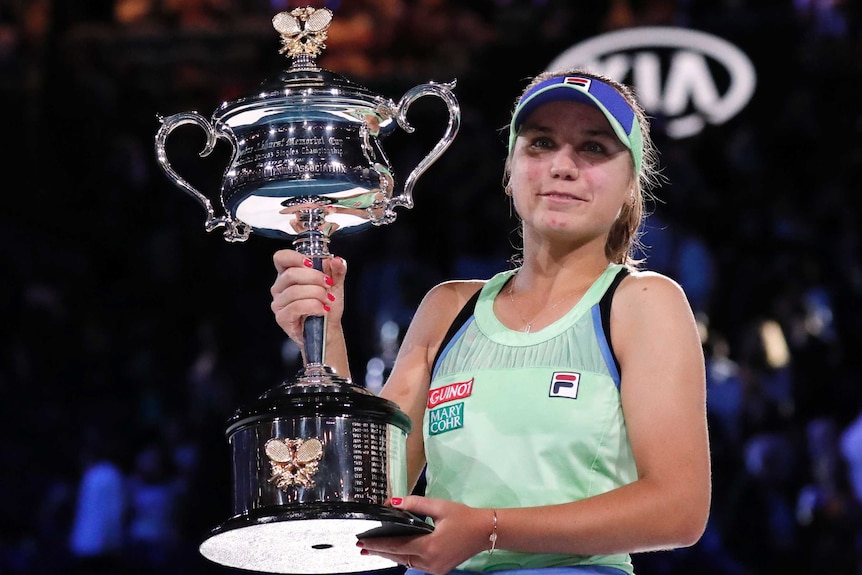 A cap-wearing tennis player holds a big trophy after winning her first Grand Slam title.