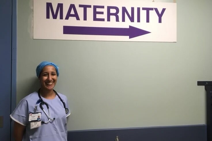 Marian smiles as she stands in theatre clothing beneath a maternity ward sign.