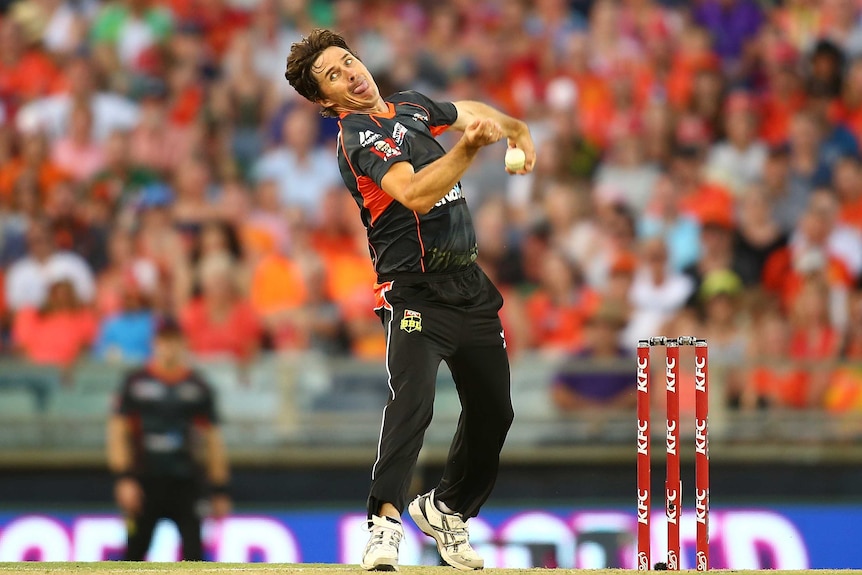 Brad Hogg bowls a cricket ball wearing a black Perth Scorchers outfit in front of a blurred crowd.