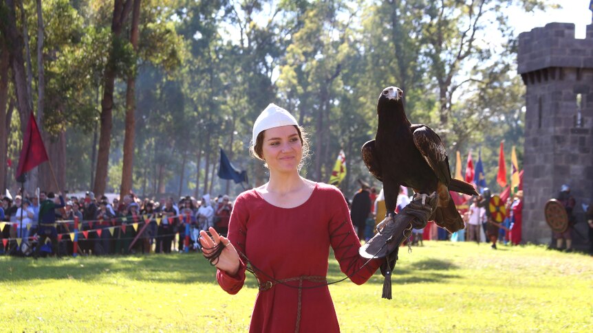 A woman in medieval dress caries an eagle in front of a castle.