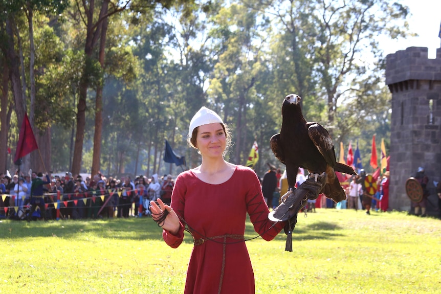 A woman in medieval dress caries an eagle in front of a castle.