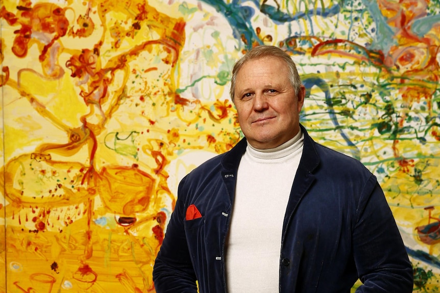 A portrait of a man in a suit standing in front of a colour john olsen artwork