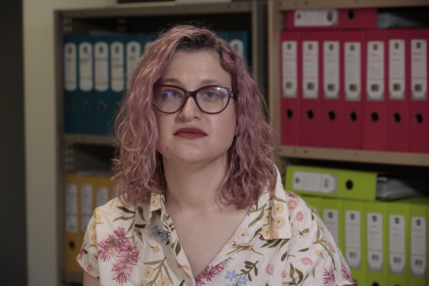 A serious woman with purple hair and glasses sits in a room with colorful binders on shelves behind her. 