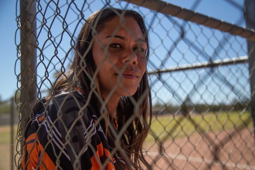 A young Indigenous woman looks at the camera through a wire fence.