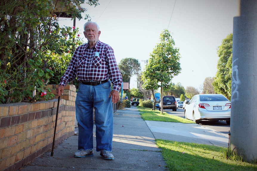 An elderly man with jeans and a checked shirt stands on a suburban street next to a brick fence.