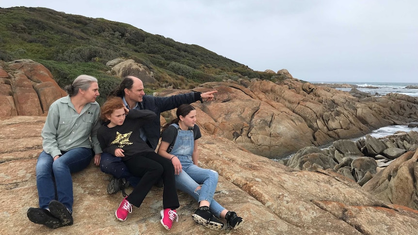 A woman, a man, and two girls sit on a rock beside the sea