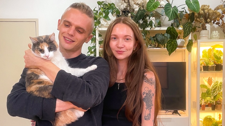Aylee and her partner smile in their home, holding their cat.