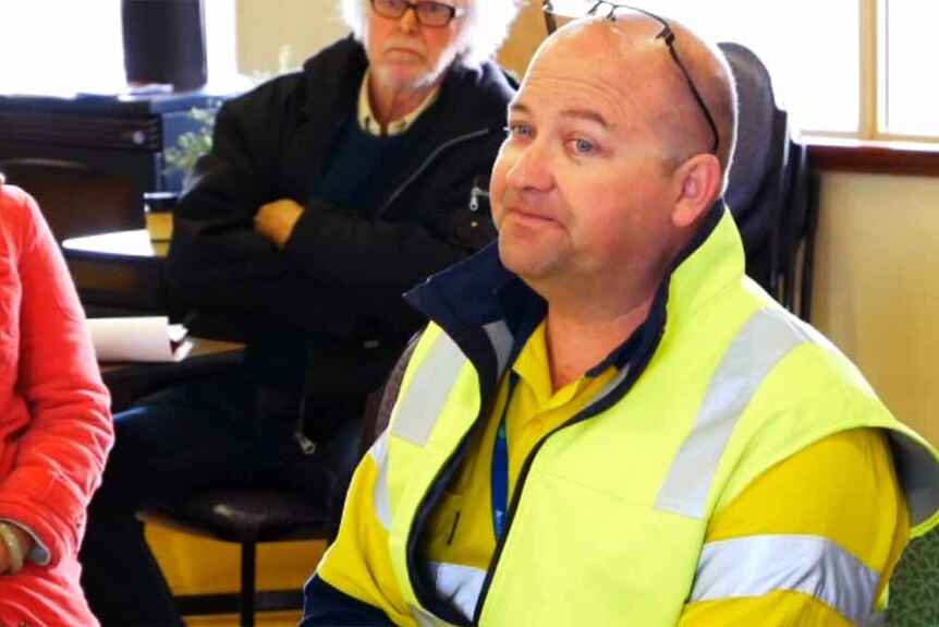 A man in a high-vis jacket sits in a room full of people.