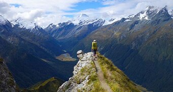 A person with backpack seen from afar at the end of long ridge, overlooking huge gully of mountains, capped in snow.