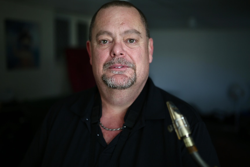 Andrew wears a black shirt, looking into the camera, while the reed of a saxophone is visible in the bottom right corner
