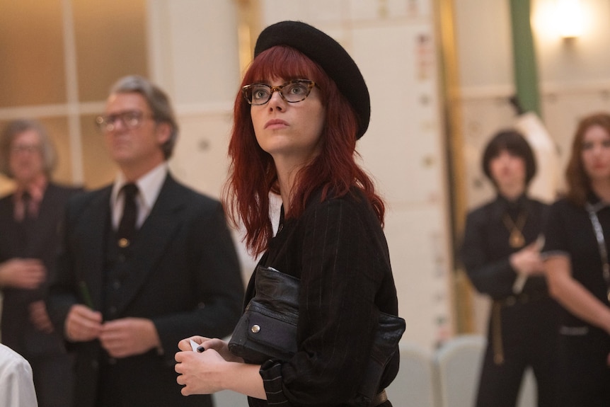 Film still of Emma Stone as Cruella with red hair and a straight fringe, dressed in a beret, with her head tilted upwards