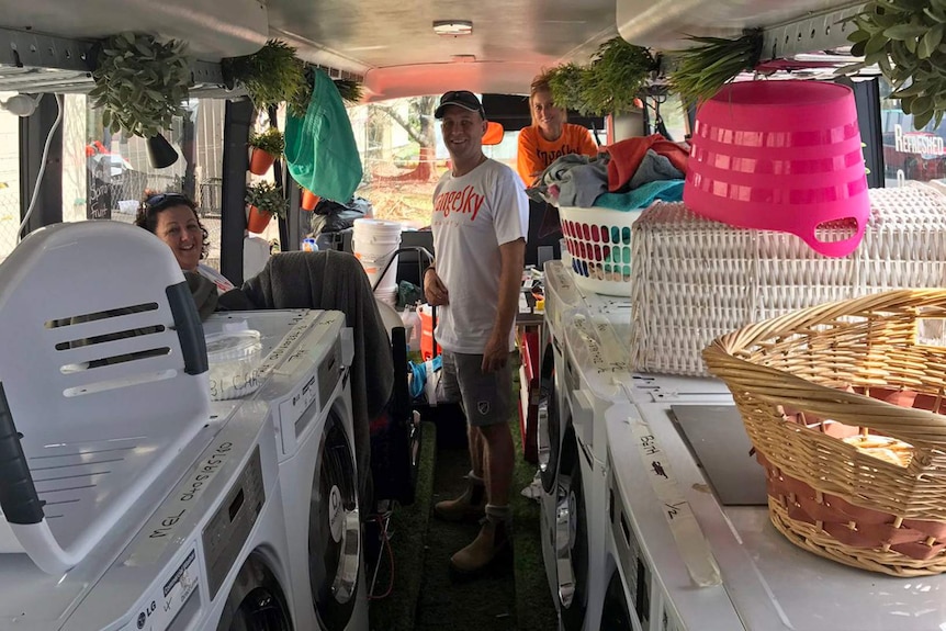 Three people stand inside a mobile laundry van surrounded by washing baskets