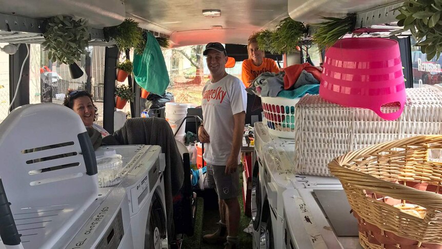 Three people stand inside a mobile laundry van surrounded by washing baskets