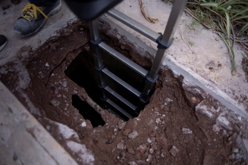 A ladder comes out of a dirt burial plot which is lowered metres below the ground, as one workers foot is captured around it.