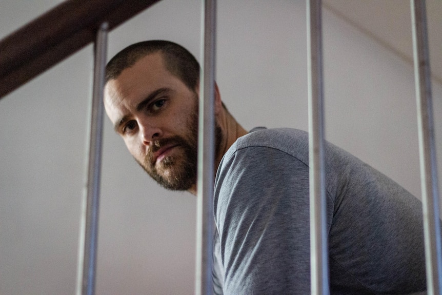 A man sits on the stairs looking at the camera behind the rungs on the stairs.