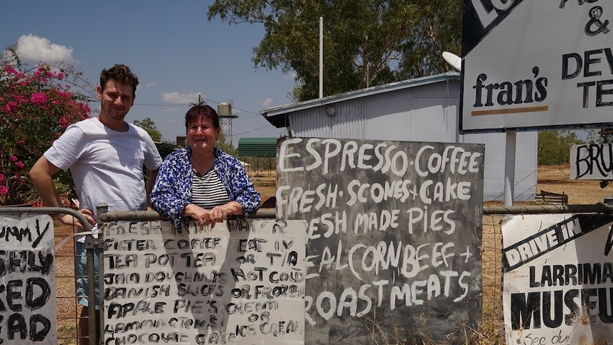 A woman leans on a waist-high fence while standing next to her adult grandson outside a shop.