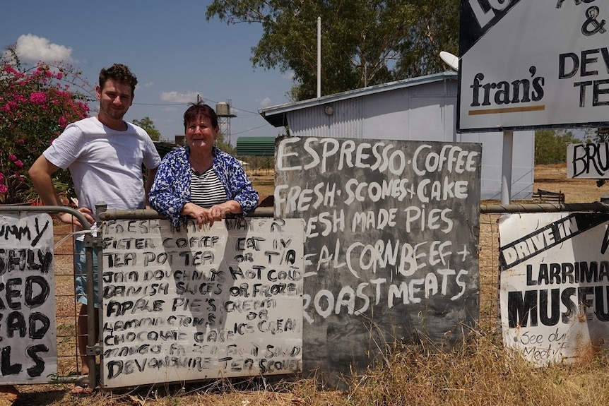 A woman leans on a waist-high fence while standing next to her adult grandson outside a shop.