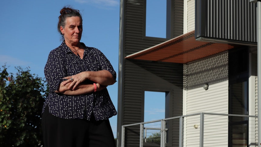 A woman stands with cross arms in front of an empty house on a suburban street.