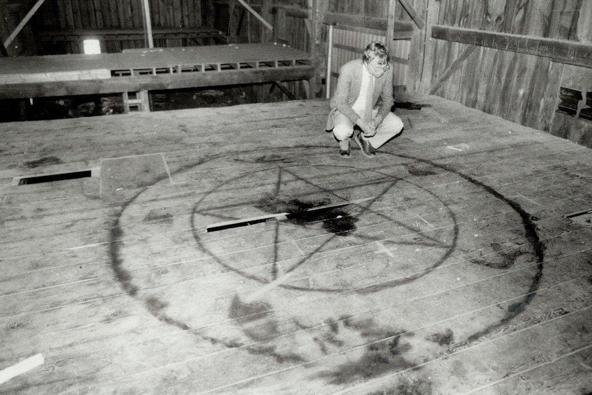 Pentagram burned onto wooden floor, man crouching beside it, in barn.