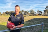 A man leans against a football ground railing while holding a football.