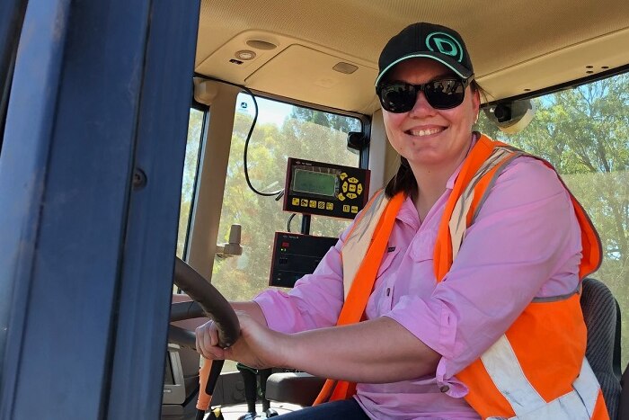 A smiling woman wearing a cap, sunglasses, hi-vis vest and pink shirt, sitting in the cabin of a tractor.