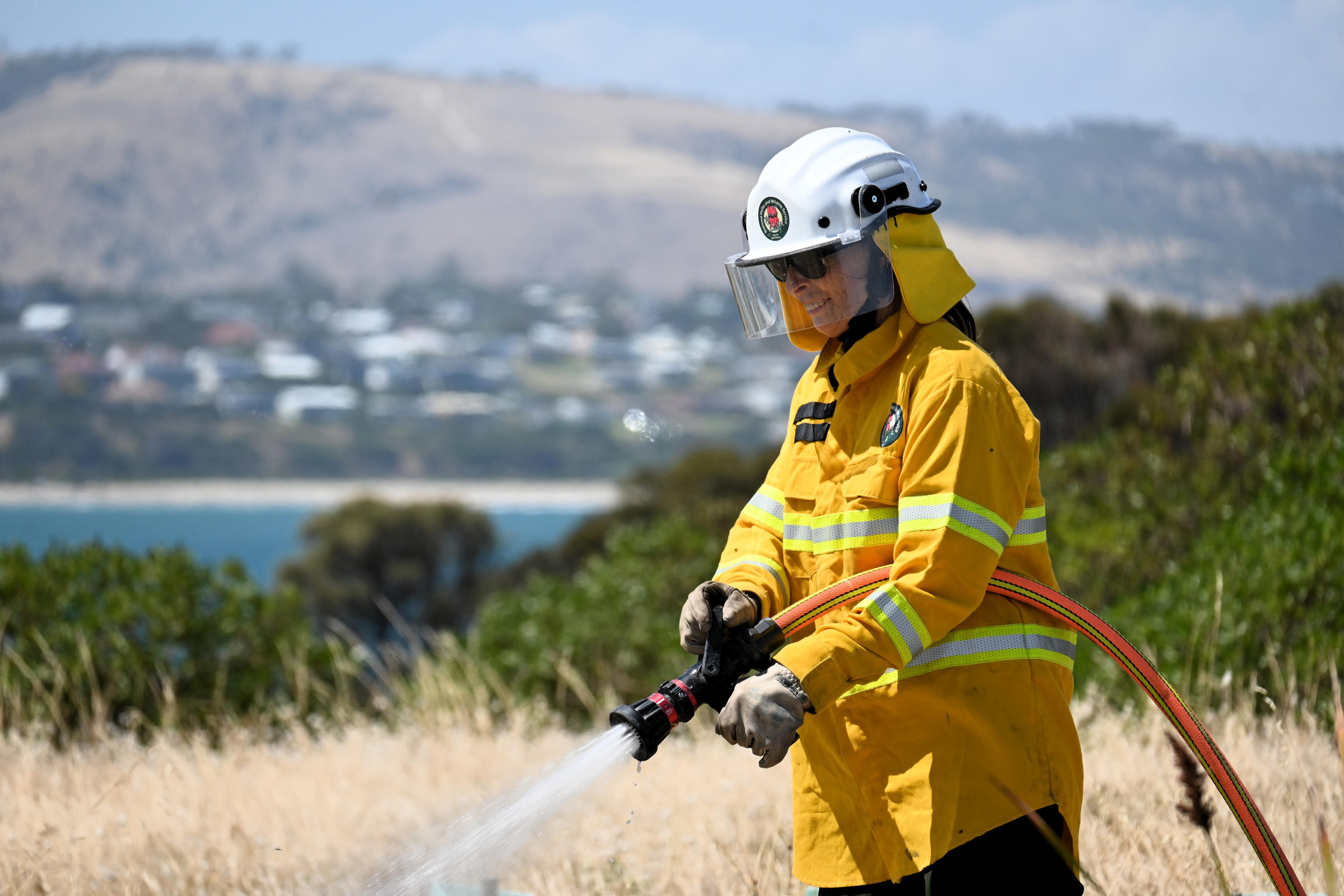 Un bombero con uniforme amarillo y casco blanco rocía el suelo con una manguera, con el mar al fondo