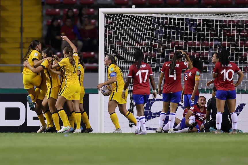 Female soccer players wearing yellow and green hug after scoring against a team in red and blue