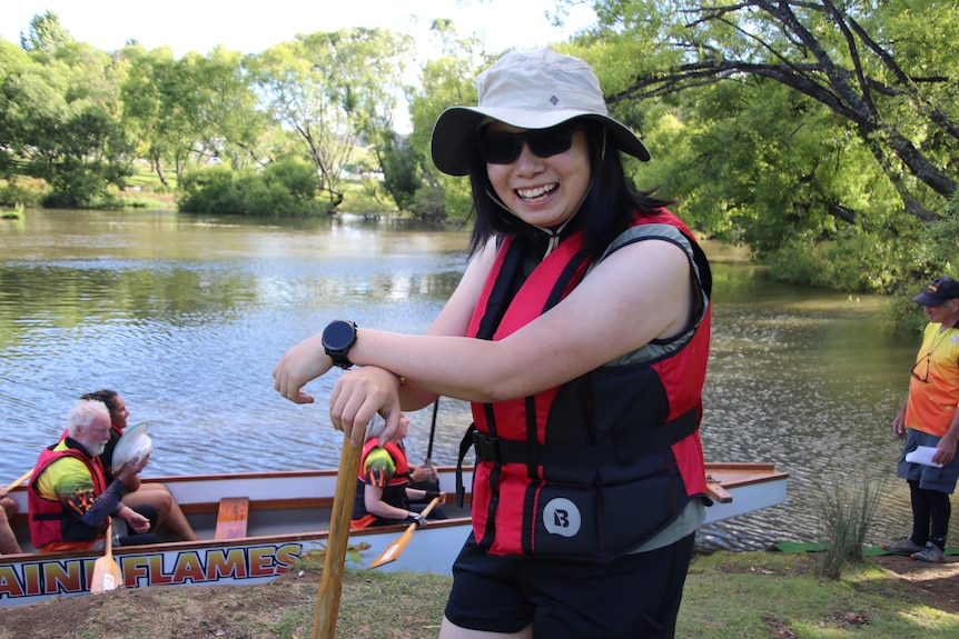 A woman wearing a wide brimmed hat leans on a dragon boat paddle and smiles.