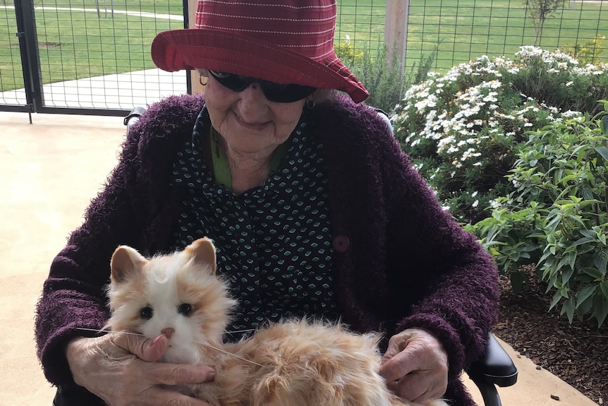 An elderly lady holds a robotic, lifelike cat on her lap.