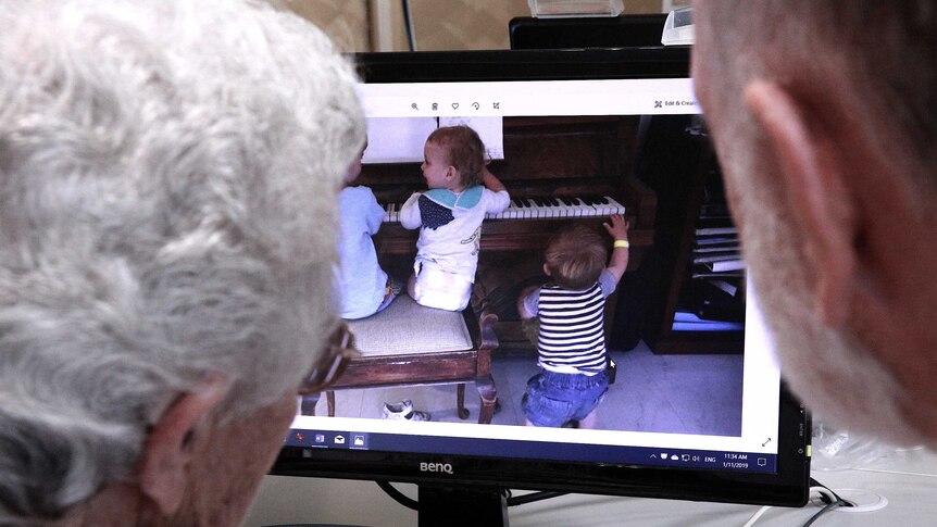 Two senior Australians look at a photo of young children playing the piano on a computer screen.