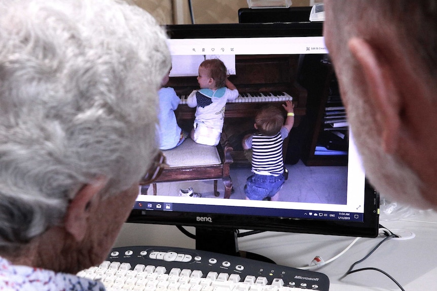 Two senior Australians look at a photo of young children playing the piano on a computer screen.