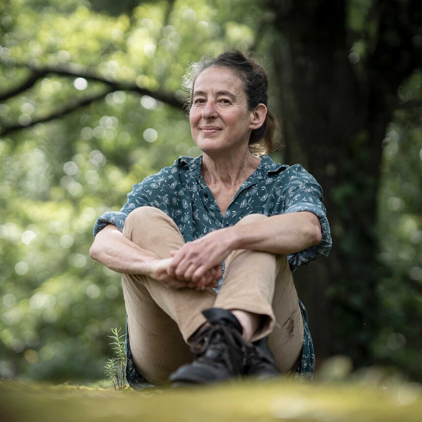 A woman in a blue blouse and light brown pants sits infront of a huge tree on the grass