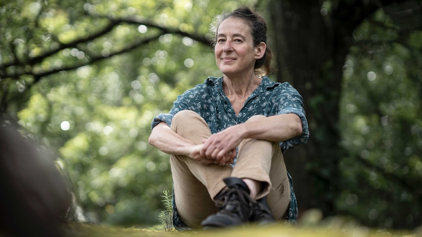 A woman in a blue blouse and light brown pants sits infront of a huge tree on the grass