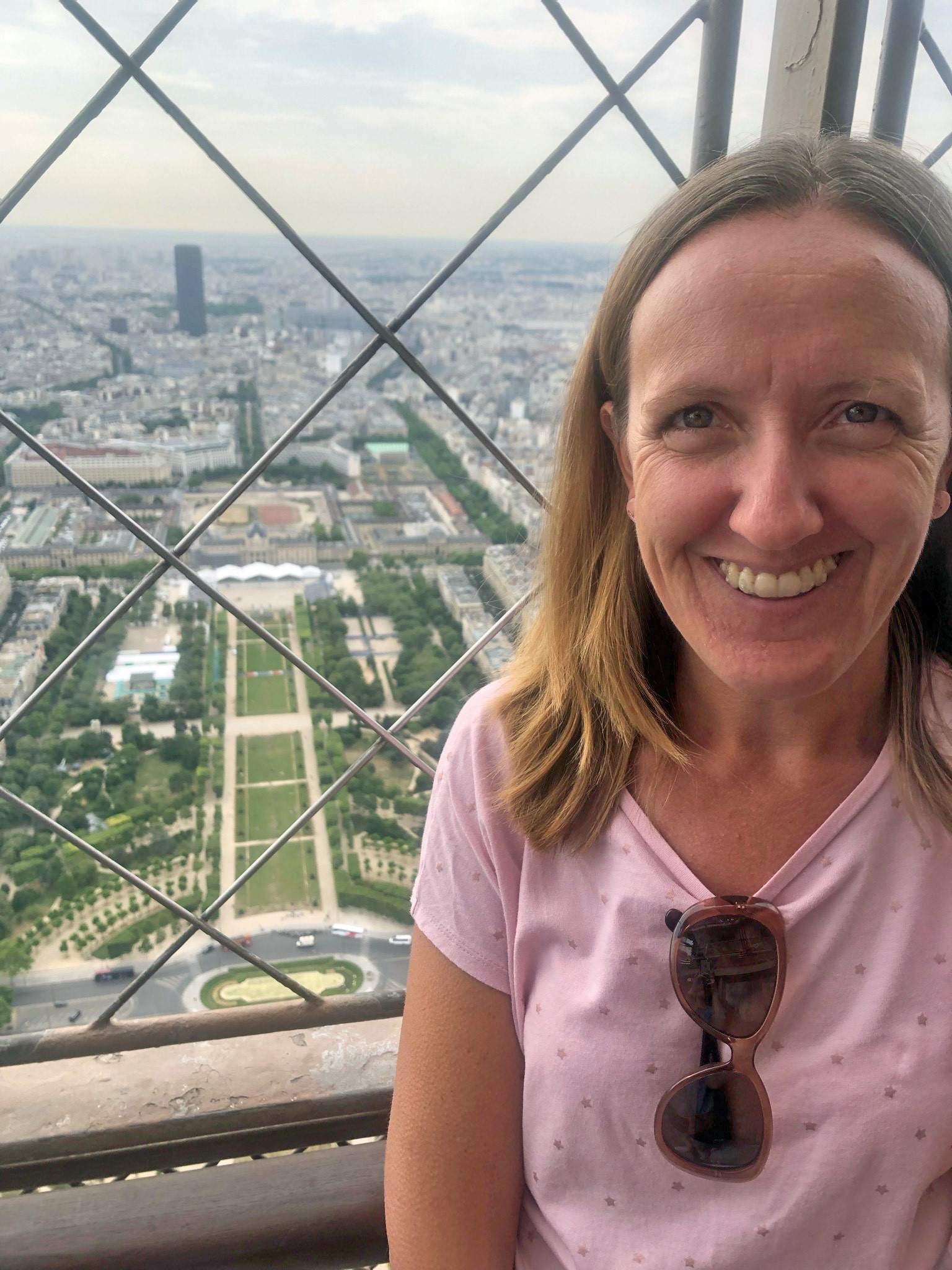 A woman with blonde hair smiles as she stands on the top of a high building, with an expansive view behind her.
