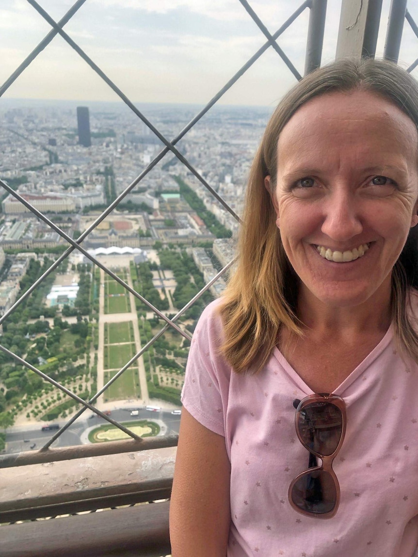 A woman with blonde hair smiles as she stands on the top of a high building, with an expansive view behind her.