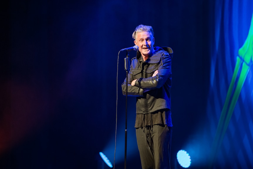 Wil Anderson, a 50-year-old man with grey hair pushed back, performs live on stage, his arms folded.. 