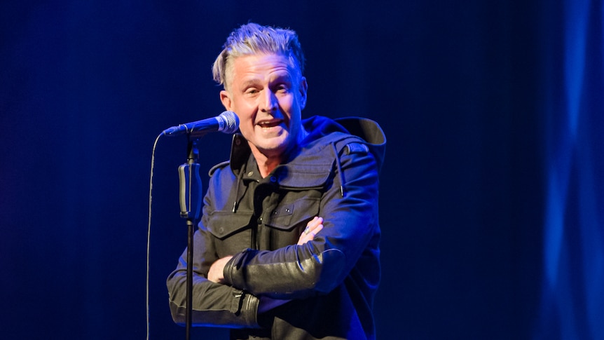 Wil Anderson, a 50-year-old man with grey hair pushed back, performs live on stage, his arms folded.. 