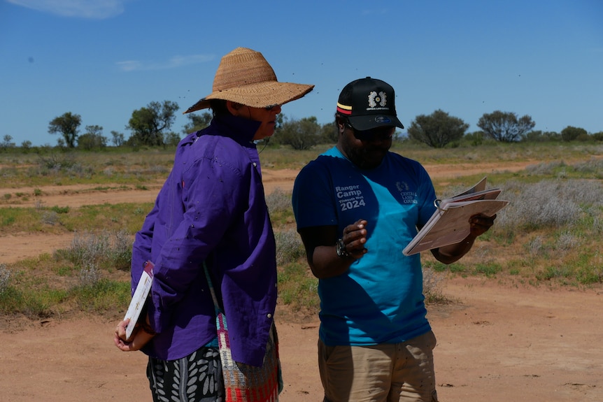 A white woman and Aboriginal man look at pages of learning documents in the desert.