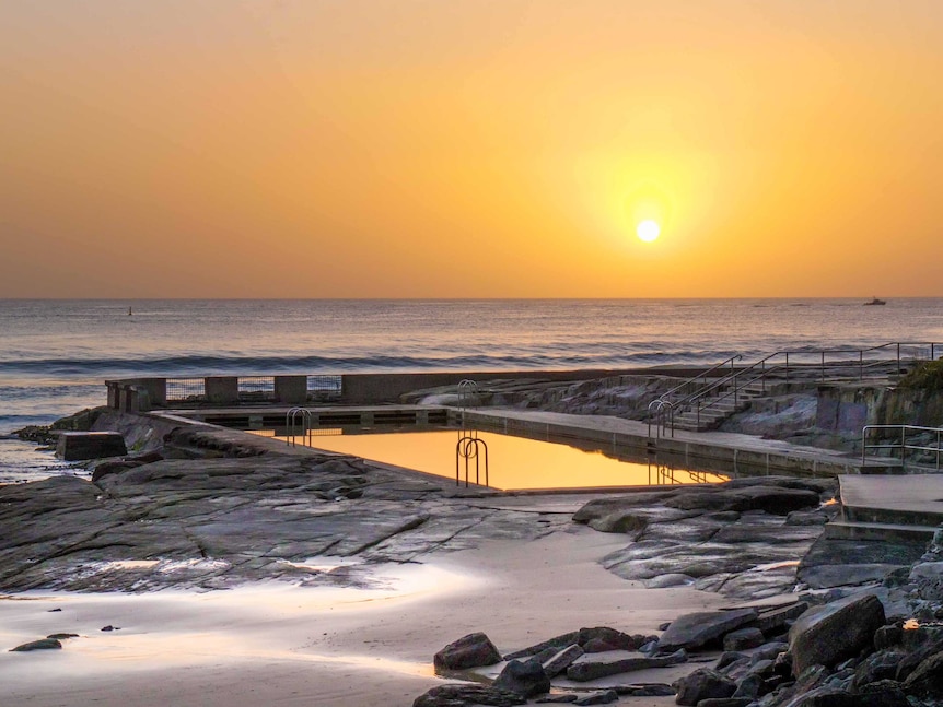 A photo of the sun rising over the Yamba ocean bath. A golden light touches the sky and the still surface of the ocean pool.