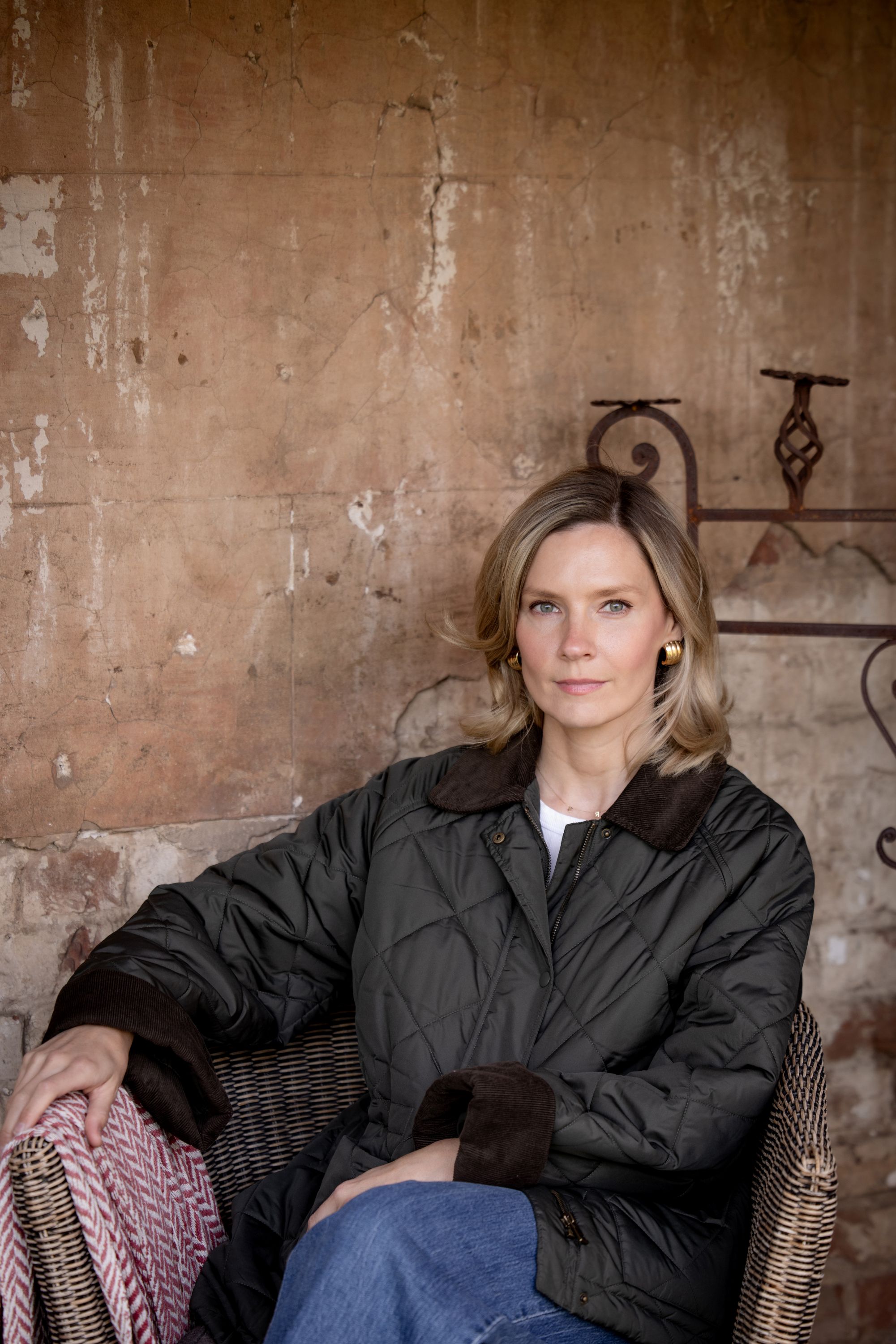 Una mujer rubia vestida con una chaqueta negra y jeans está sentada con las piernas cruzadas frente a una pared exterior enlucida.