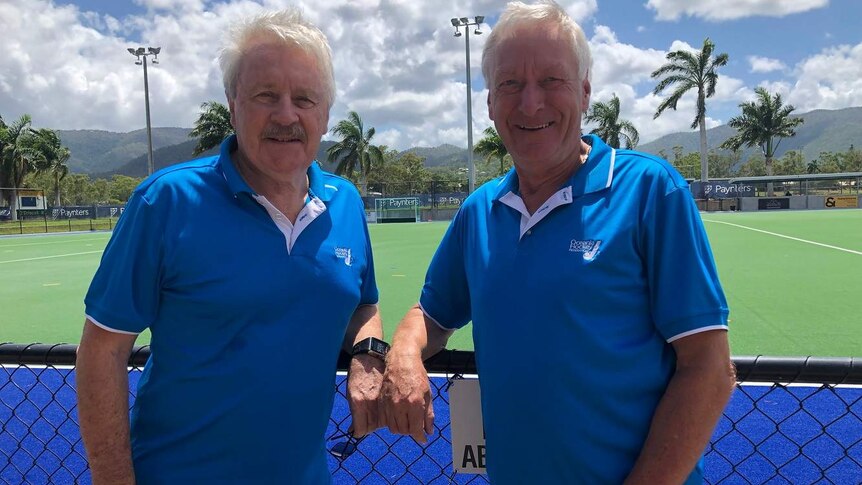 Two men with grey hair wear blue shirts and stand in front of a green hockey field.