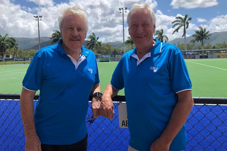 Two men with grey hair wear blue shirts and stand in front of a green hockey field.