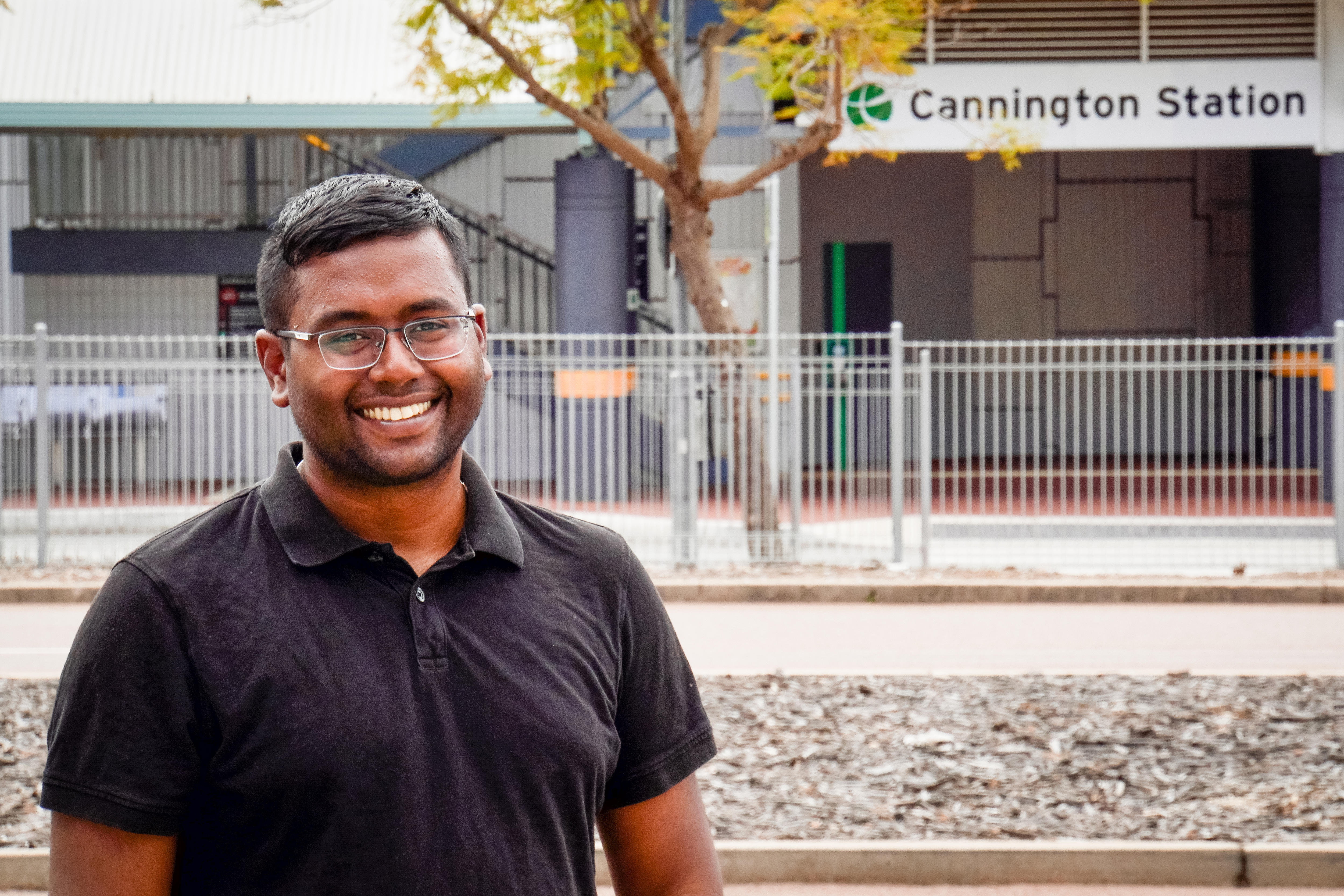 A smiling, tanned man wearing glasses and a black polo shirt. Cannington train station sign in the background.