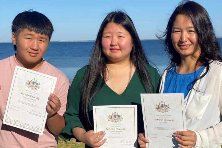 Boy, girl and woman holding citizenship certificates and looking at camera, beach in background