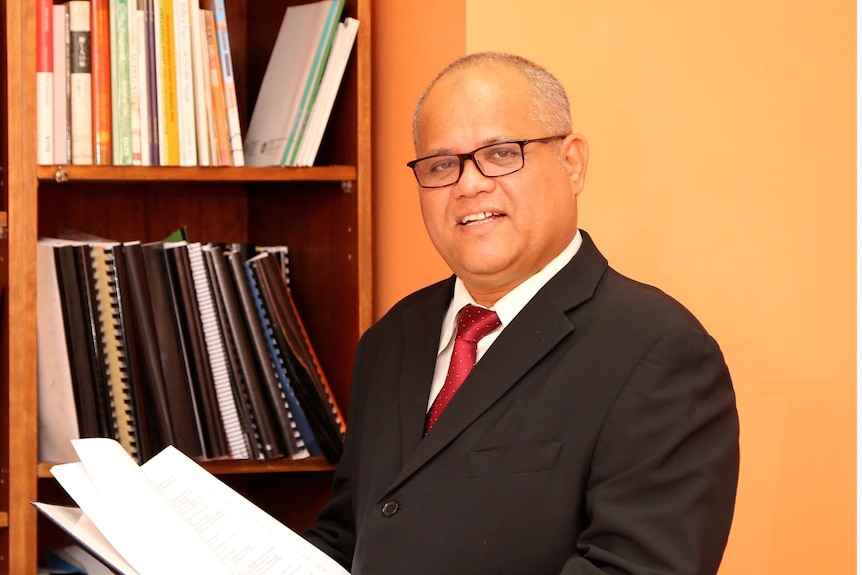 A bald man in a suit standing in front of a suitcase.